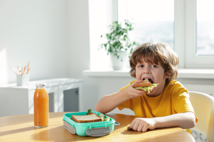 little boy eating a sandwich at a table with his lunch box and a juice in front of him. 