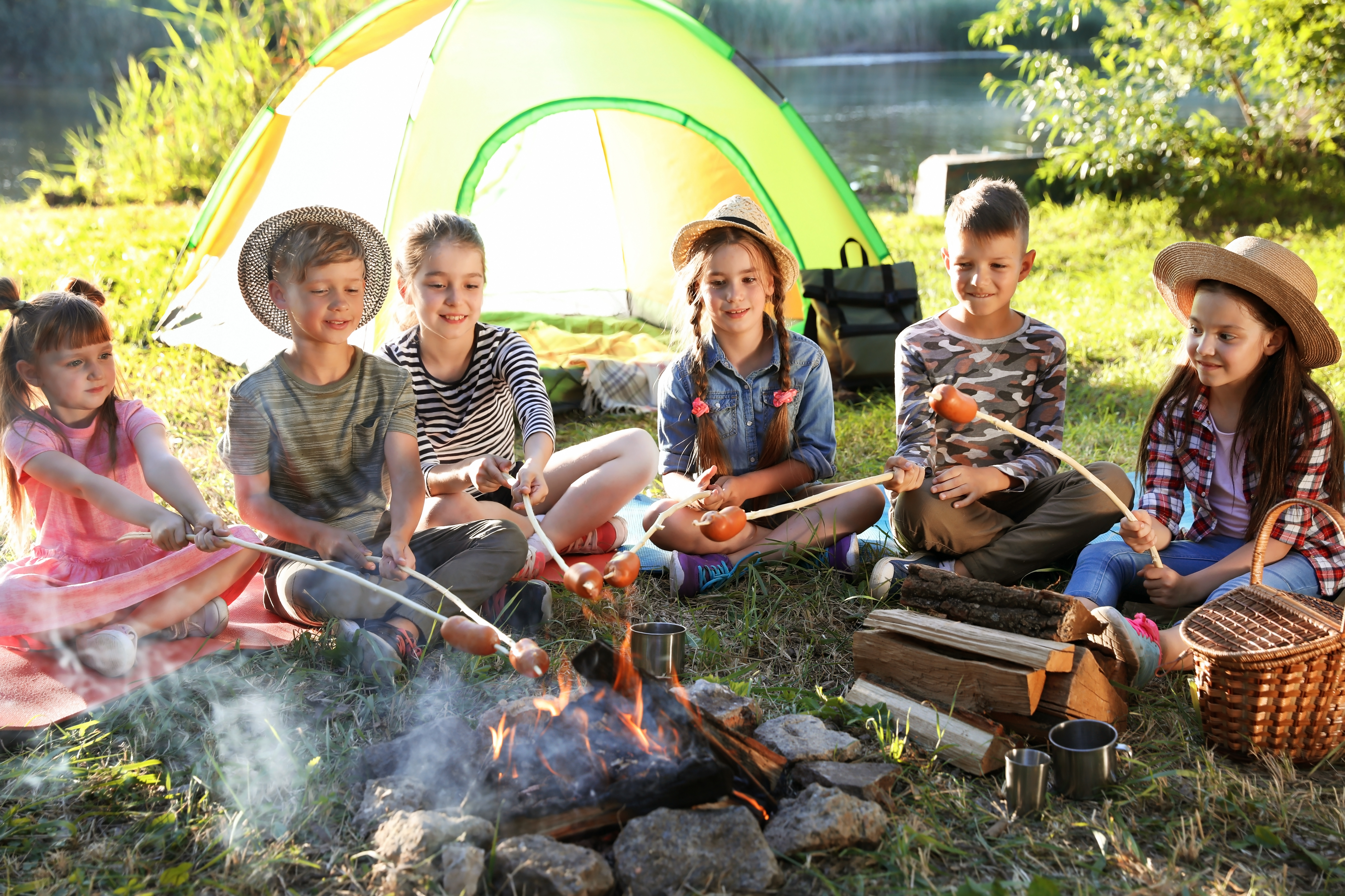 kids sitting on the ground at a campsite in front of a fire roasting sausages