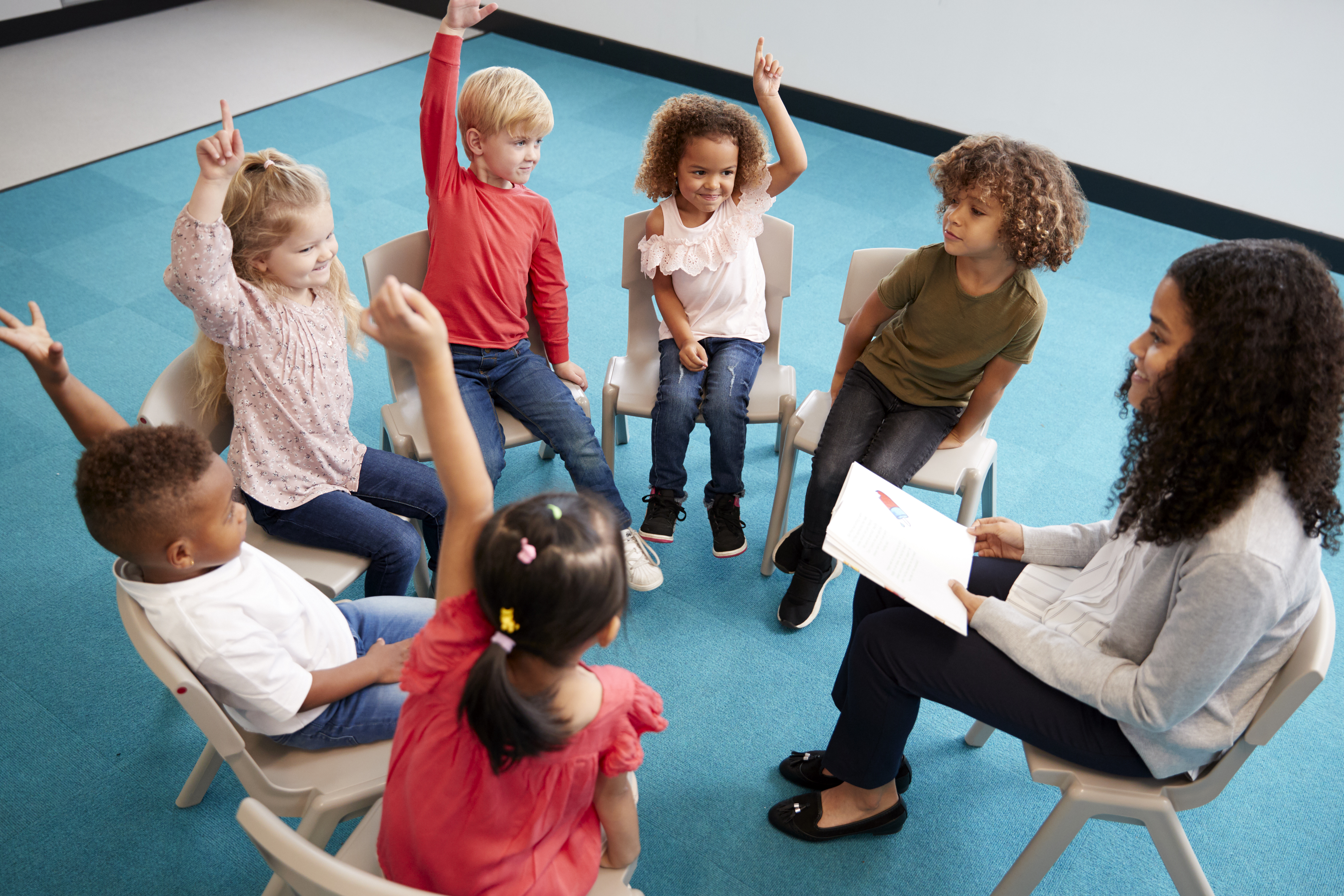 female teacher and very young students sitting in a circle in chairs with their hands raised